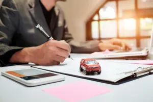 Person in suit writing on document, toy car, calculator, and laptop on a desk.