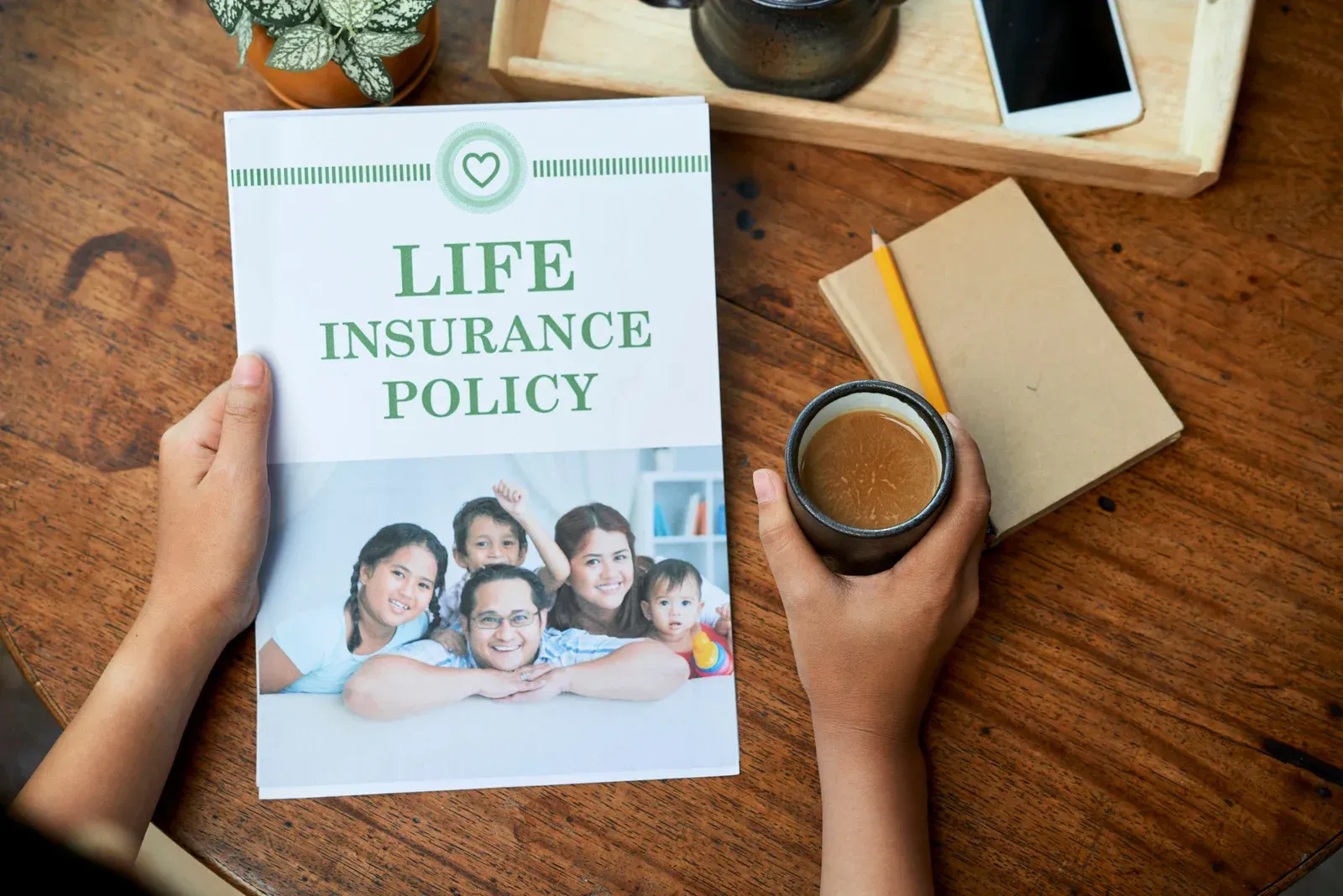 Person holding a life insurance policy, family photo on the cover, sitting at a table with coffee and a notebook.