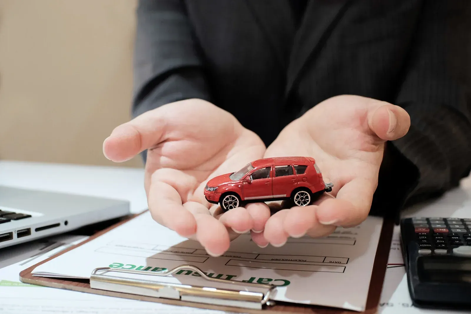 Person in suit holding a red toy car over car insurance paperwork; calculator and laptop present.