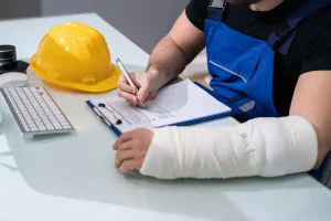 Construction worker with arm in cast, writing on clipboard at desk. Hard hat and keyboard nearby.