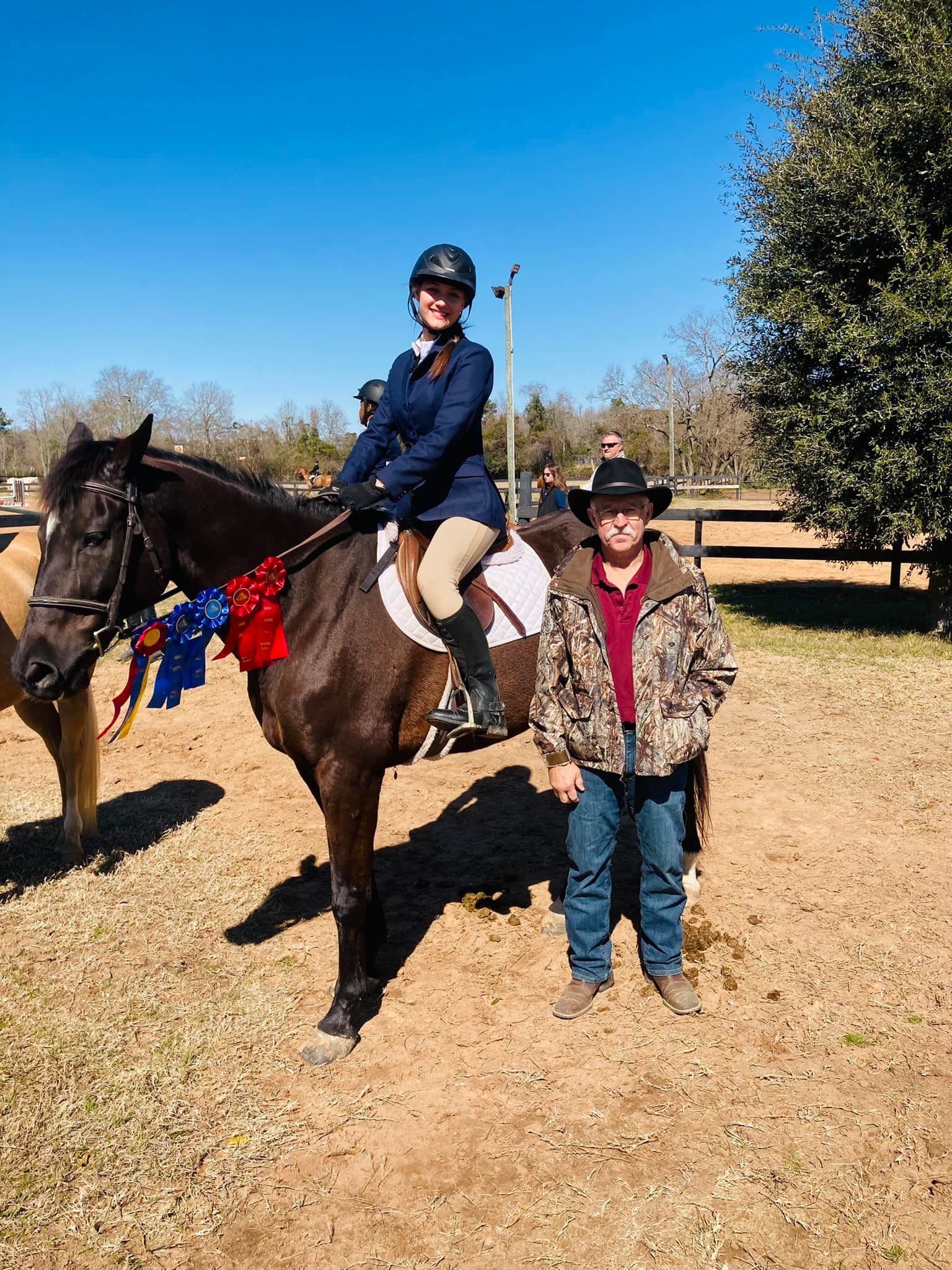 A rider in equestrian attire sits atop a dark horse with show ribbons, posing with a person in camo outdoors.
