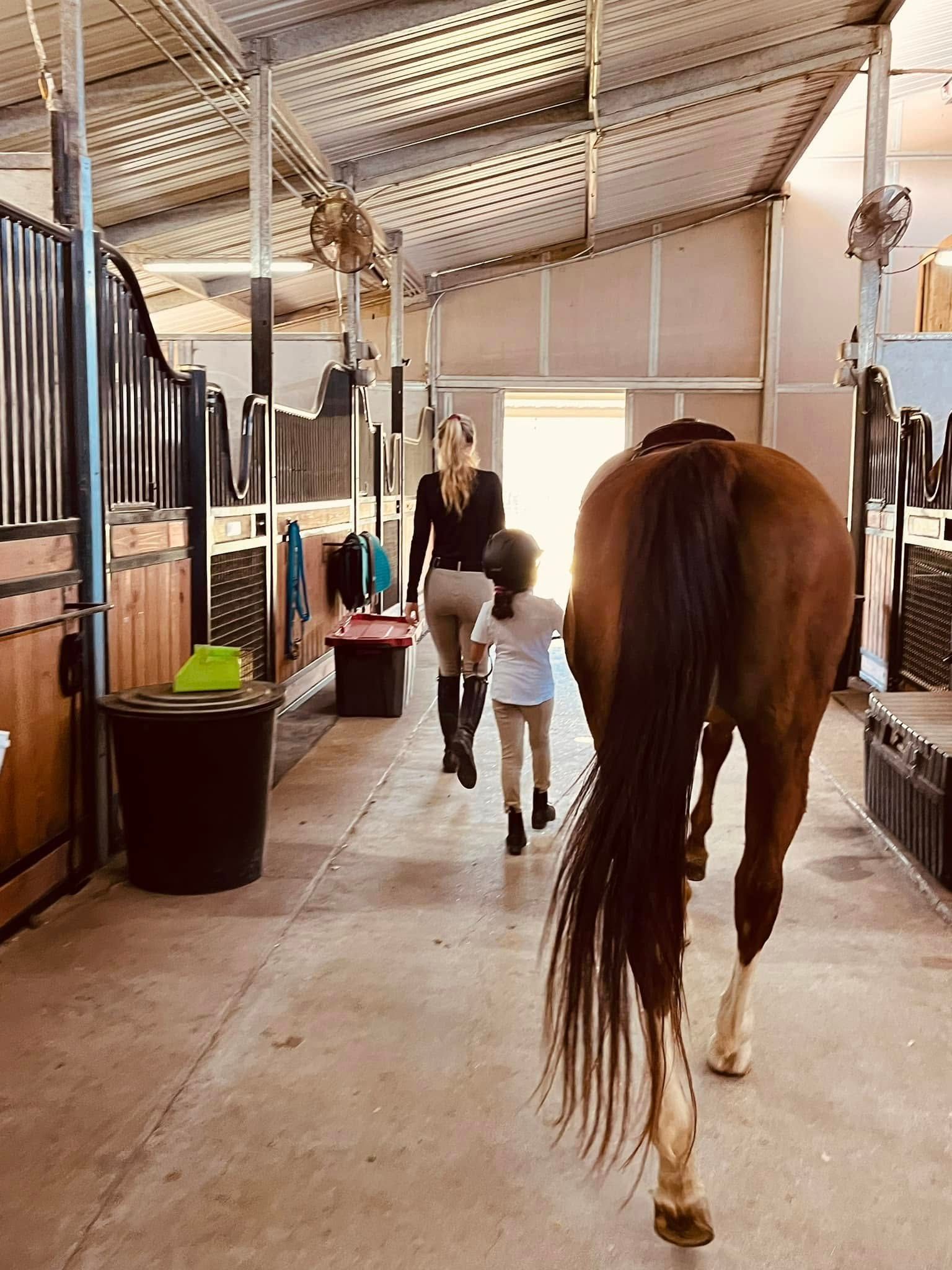 A person and child walking alongside a brown horse down a stable aisle toward a bright doorway.