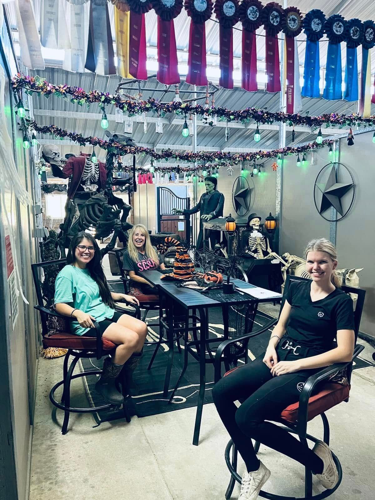 Three people sit around a black table in a room decorated for Halloween with hanging ribbons, skeletons, and lights.