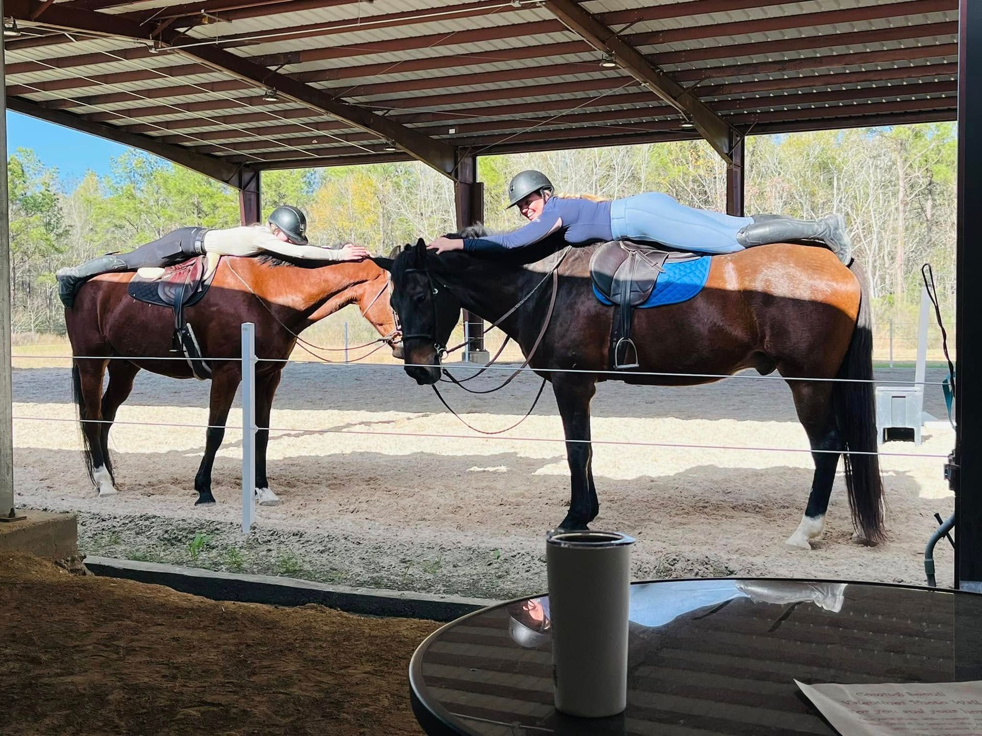 Two riders lie prone on the backs of their horses in a covered outdoor arena.