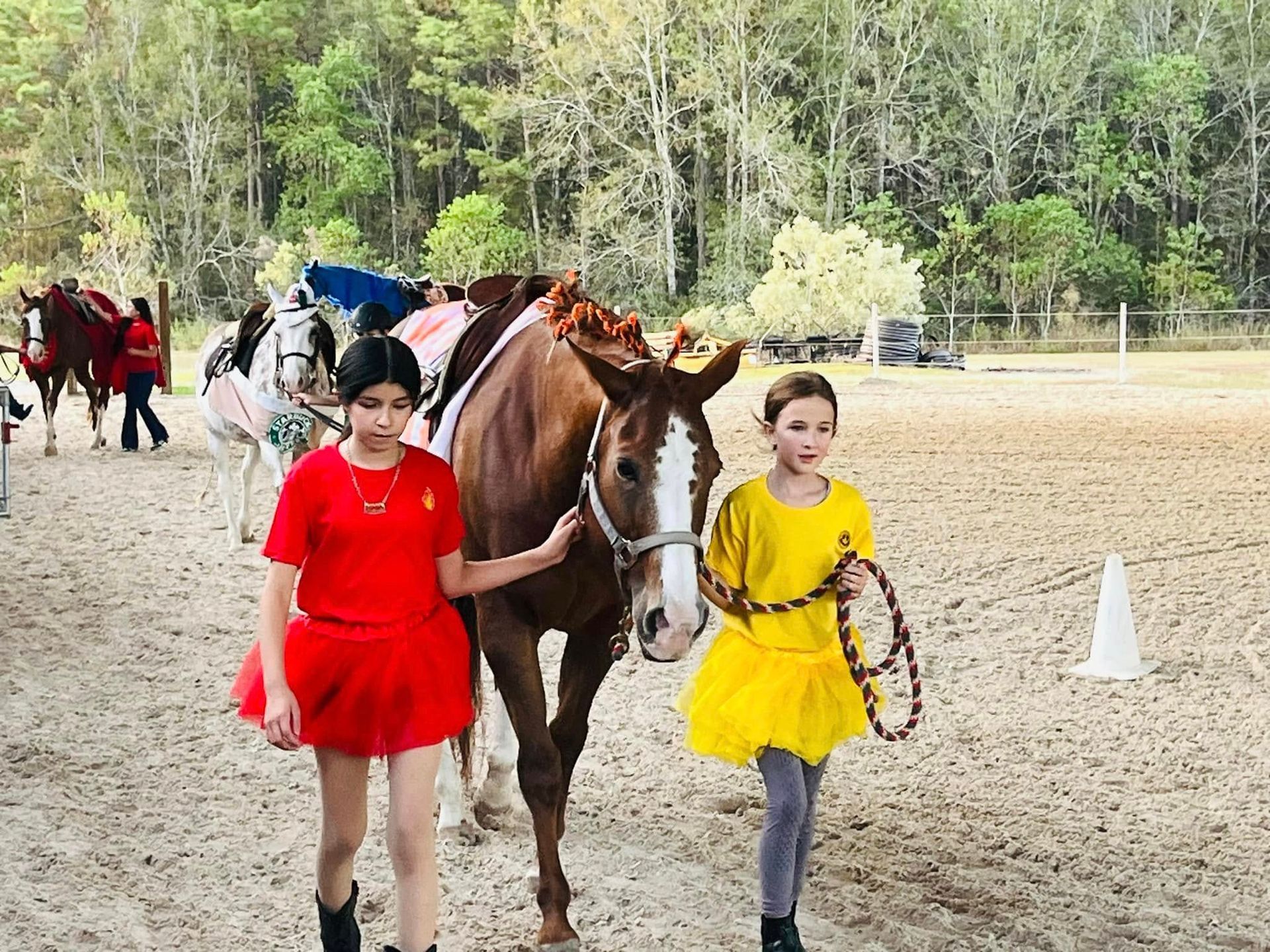 Two children in red and yellow outfits lead a brown horse across a dirt riding ring with trees in the background.