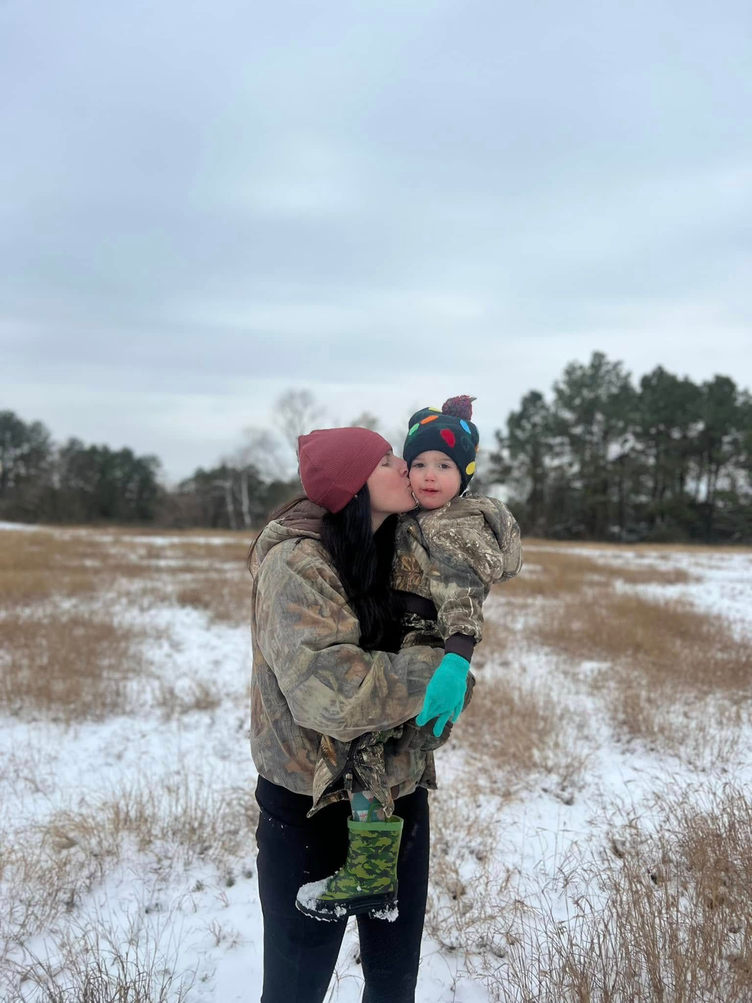 A person in a camouflage jacket kisses a child wearing a patterned hat in a snowy, open field under a cloudy sky.