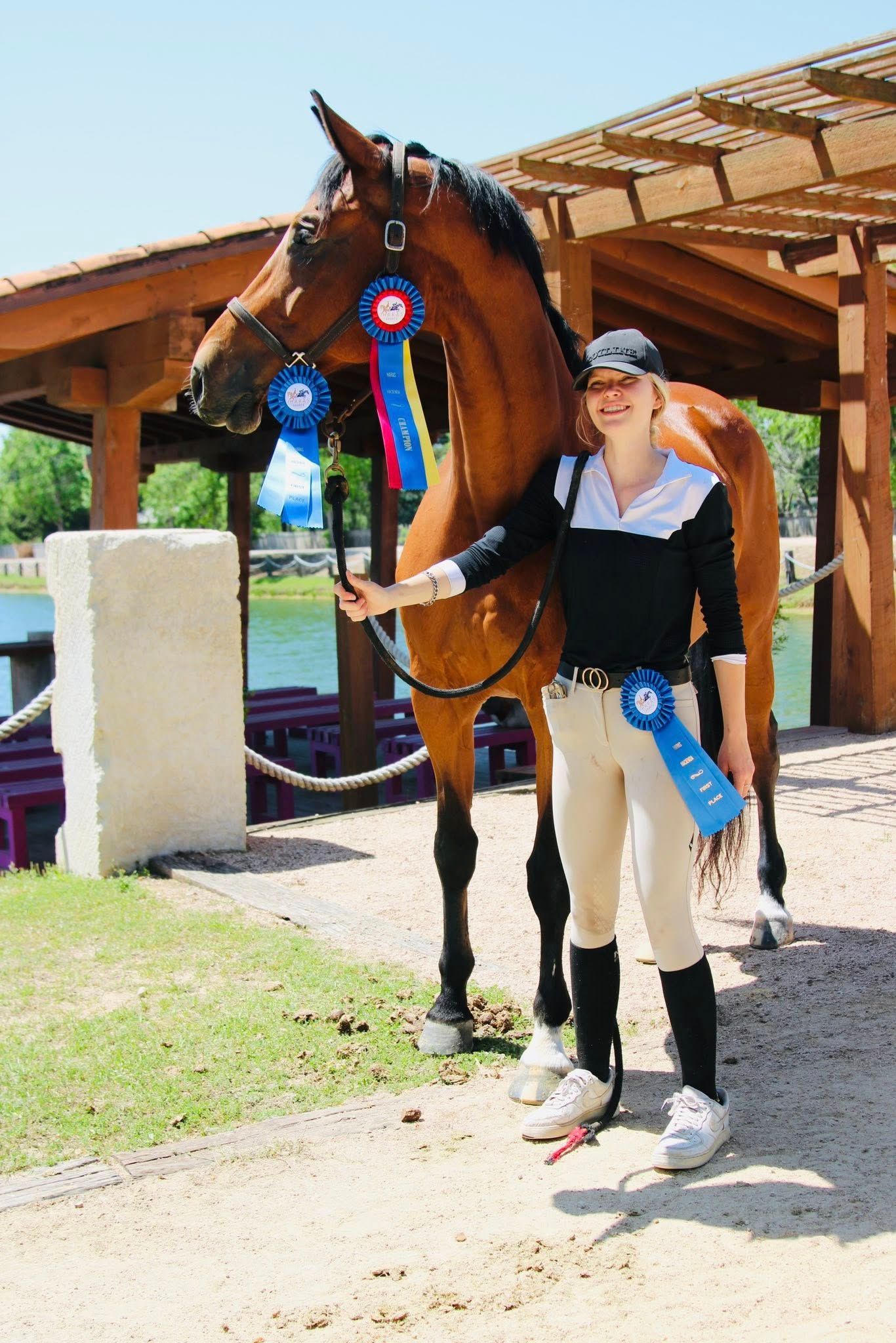 A person in equestrian gear stands beside a brown horse wearing blue award ribbons under a wooden shelter near water.