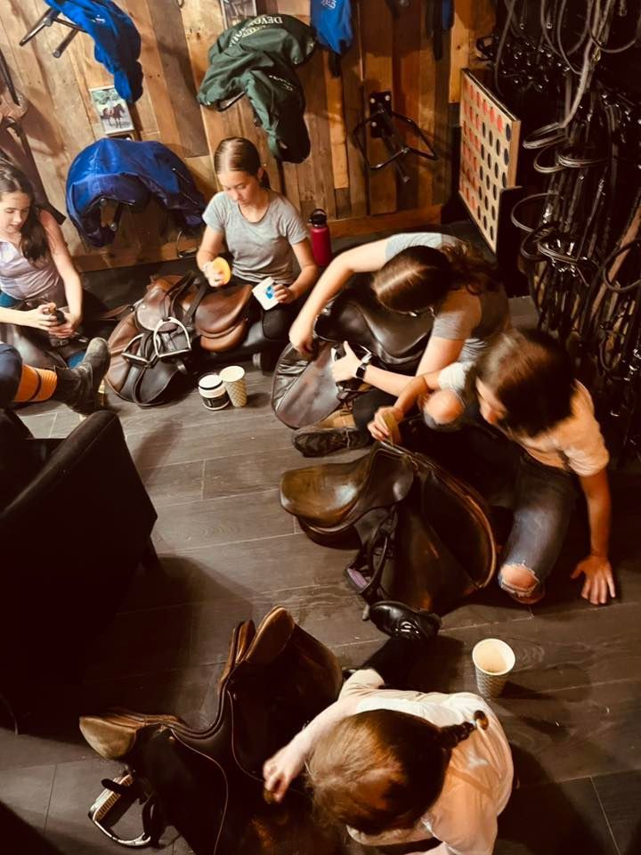 Several people sit on a wooden floor in a barn, cleaning and maintaining leather horse saddles.