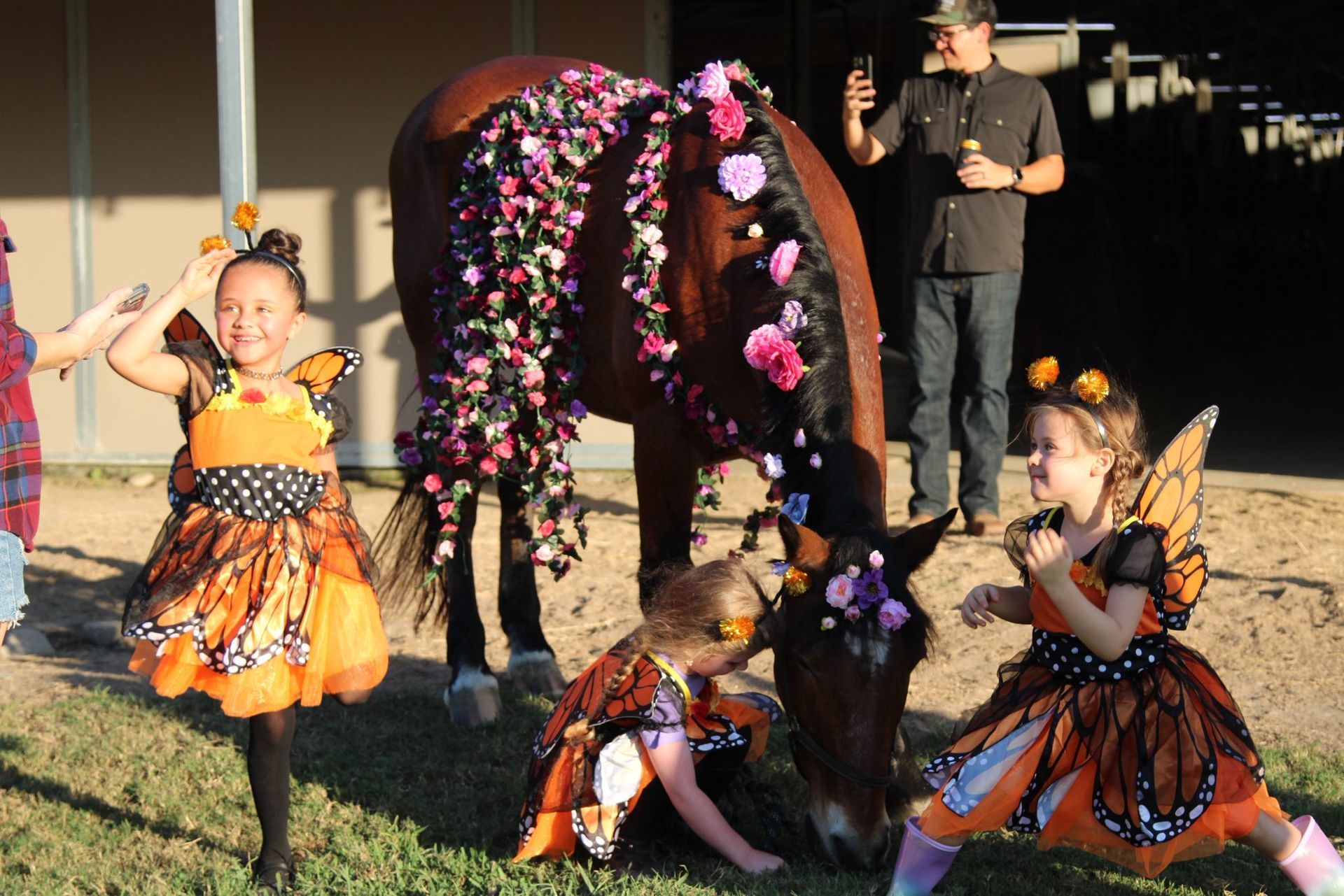 Three children in butterfly costumes interact with a flower-adorned horse at a stable while a person films them.