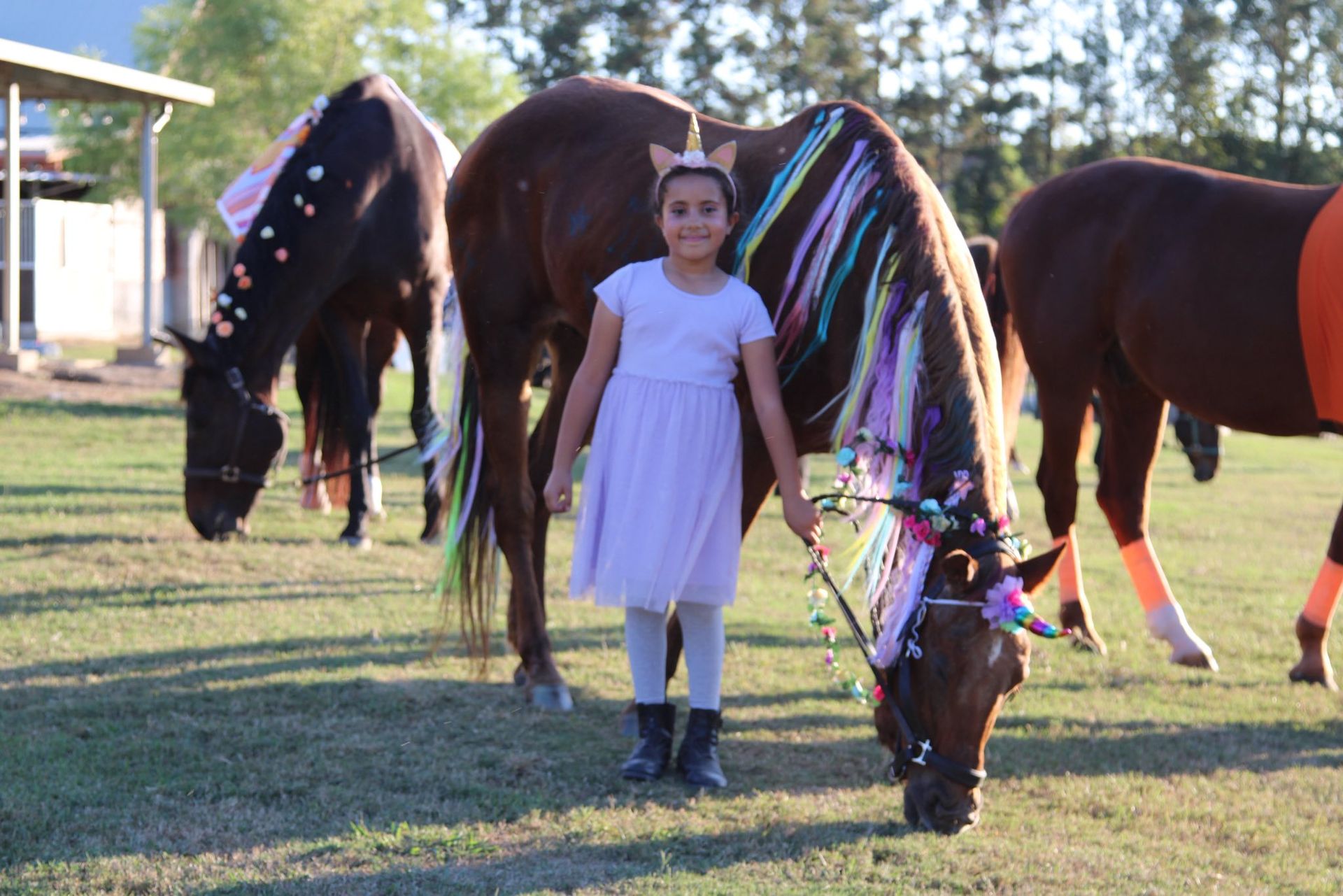 A person wearing a unicorn headband and a light-colored dress stands between two horses adorned with ribbons and flowers.