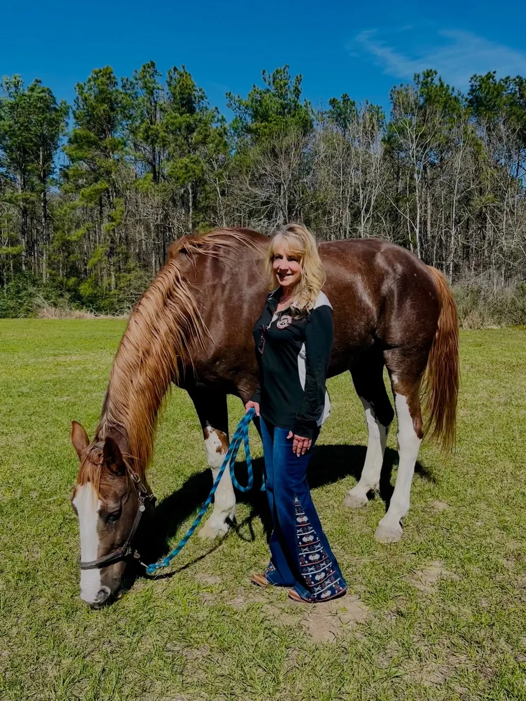 A woman stands in a grassy field holding the lead rope of a brown and white horse that is grazing.