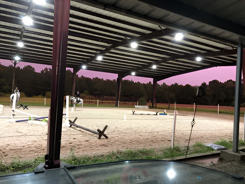 A covered horse arena at dusk with riders on horseback practicing jumps in a sand-filled ring under bright overhead lights.