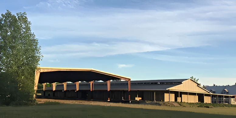 A large, open-sided metal structure with a prominent roof overhang stands in a grassy field under a blue sky.