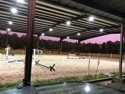 Two equestrians ride horses inside a large, covered outdoor arena at dusk, with artificial lights illuminating the ring.
