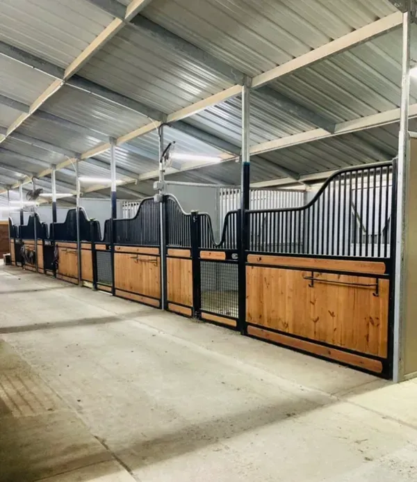 A row of horse stalls with wooden lower walls and black metal upper bars inside a modern, covered stable building.