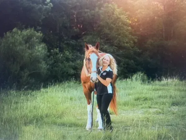 A woman standing in a grassy field, smiling while holding the reins of a chestnut horse with a white blaze on its face.