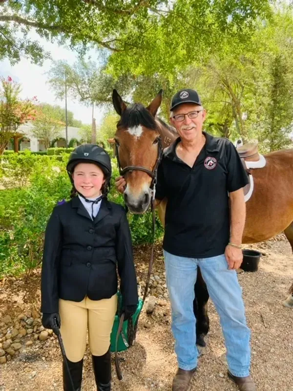 A person in equestrian attire and a person in a black polo shirt stand outdoors next to a brown horse.