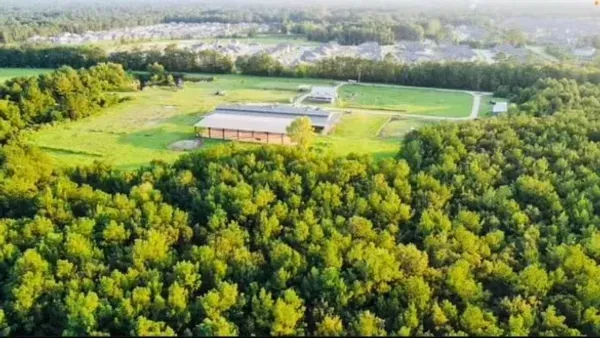 An aerial view of a large building in a clearing, surrounded by dense forest with a residential neighborhood in the distance.