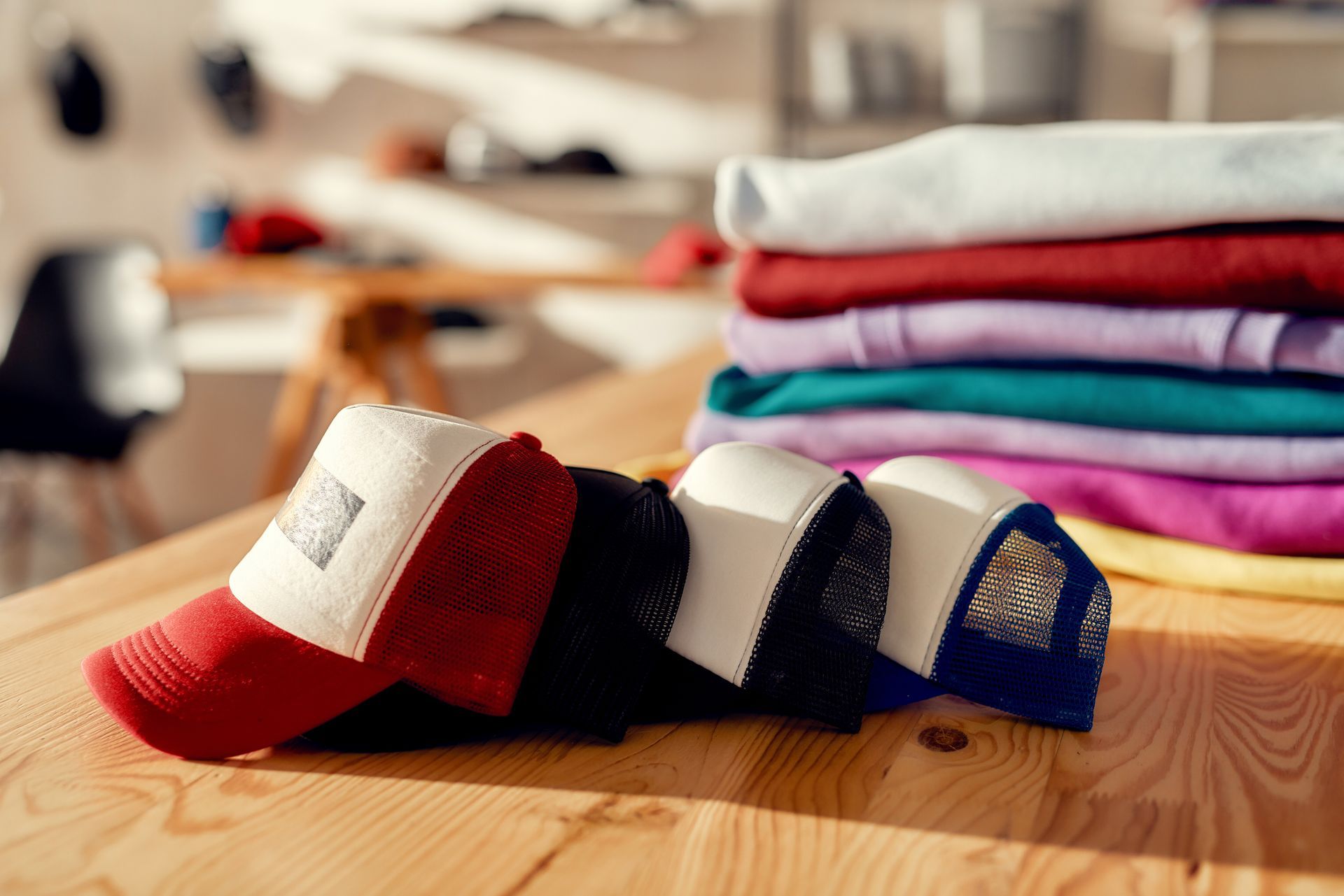 Custom apparel store display with stacked hats and folded t-shirts on wooden table in print shop.
