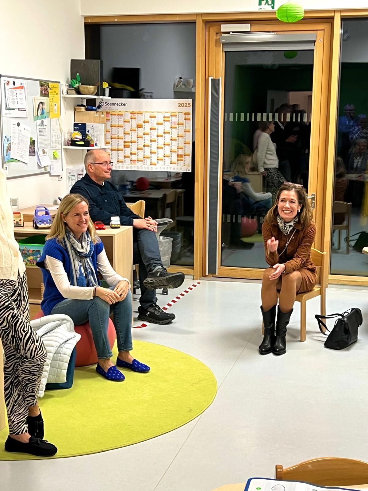 Menschen sitzen in einem Raum, in der Mitte steht eine lachende Frau. Auf dem Boden liegt ein grüner runder Teppich.