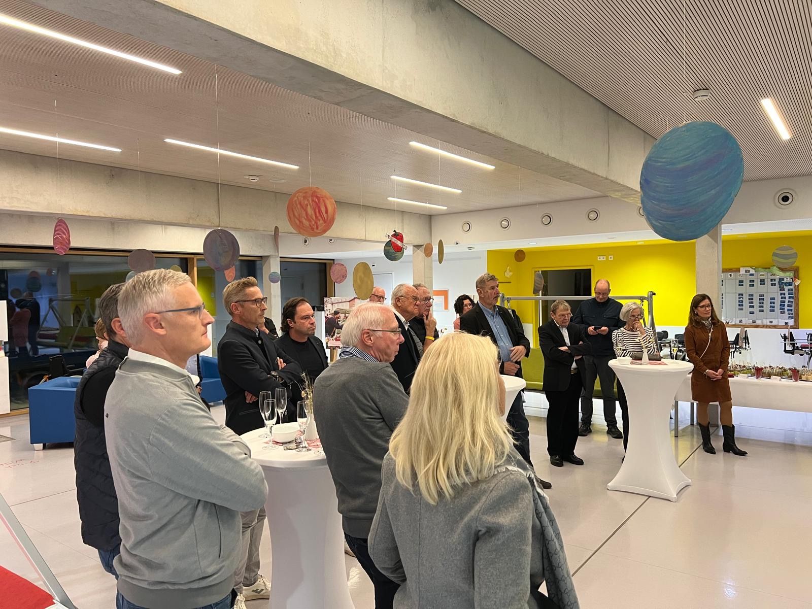 Menschen haben sich in einem hell erleuchteten Raum mit farbenfroher Dekoration versammelt, einige hören einem Redner zu.