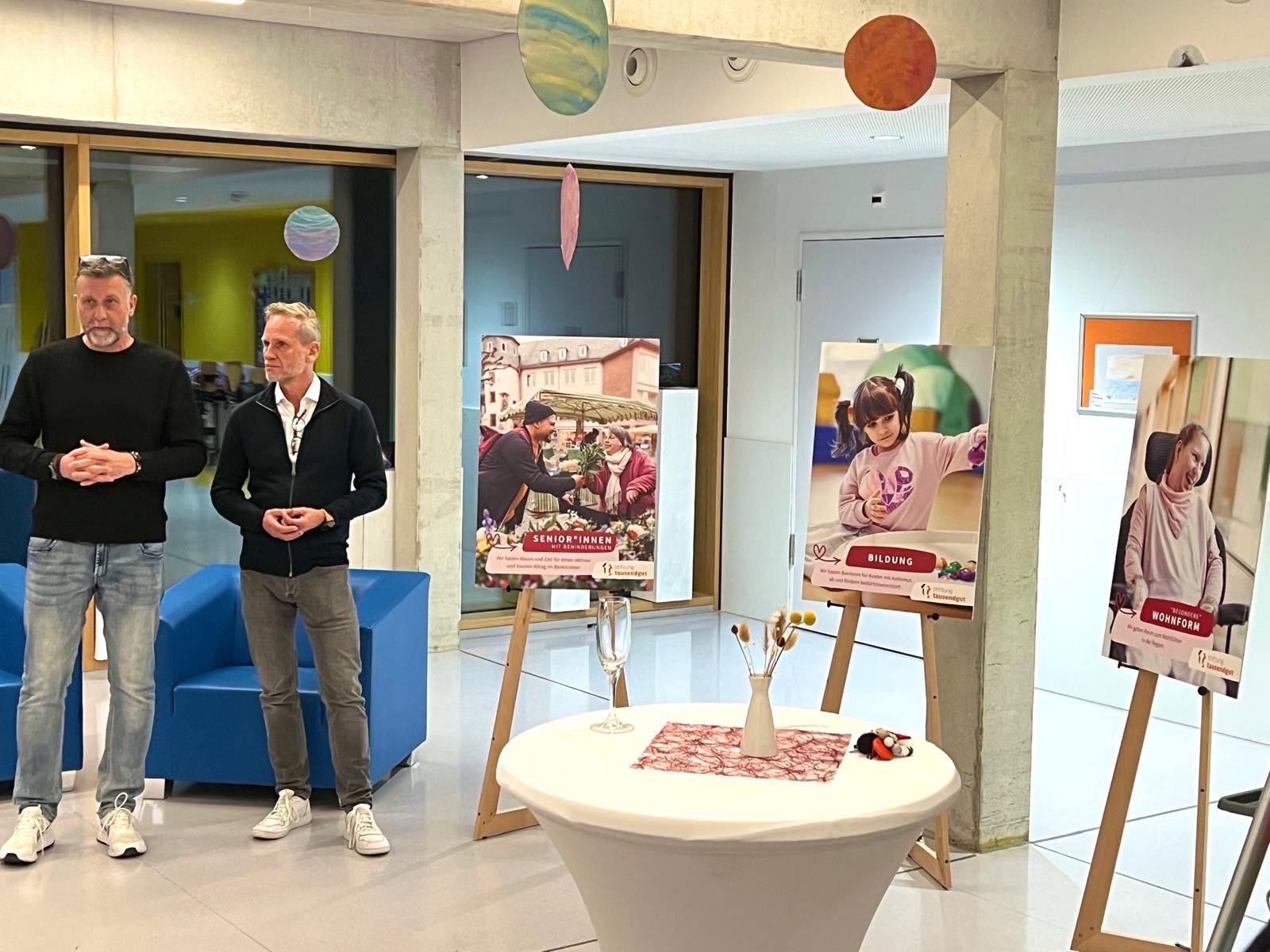 Zwei Männer stehen bei einer Veranstaltung vor Plakaten. Über einem kleinen Tisch hängen Luftballons.