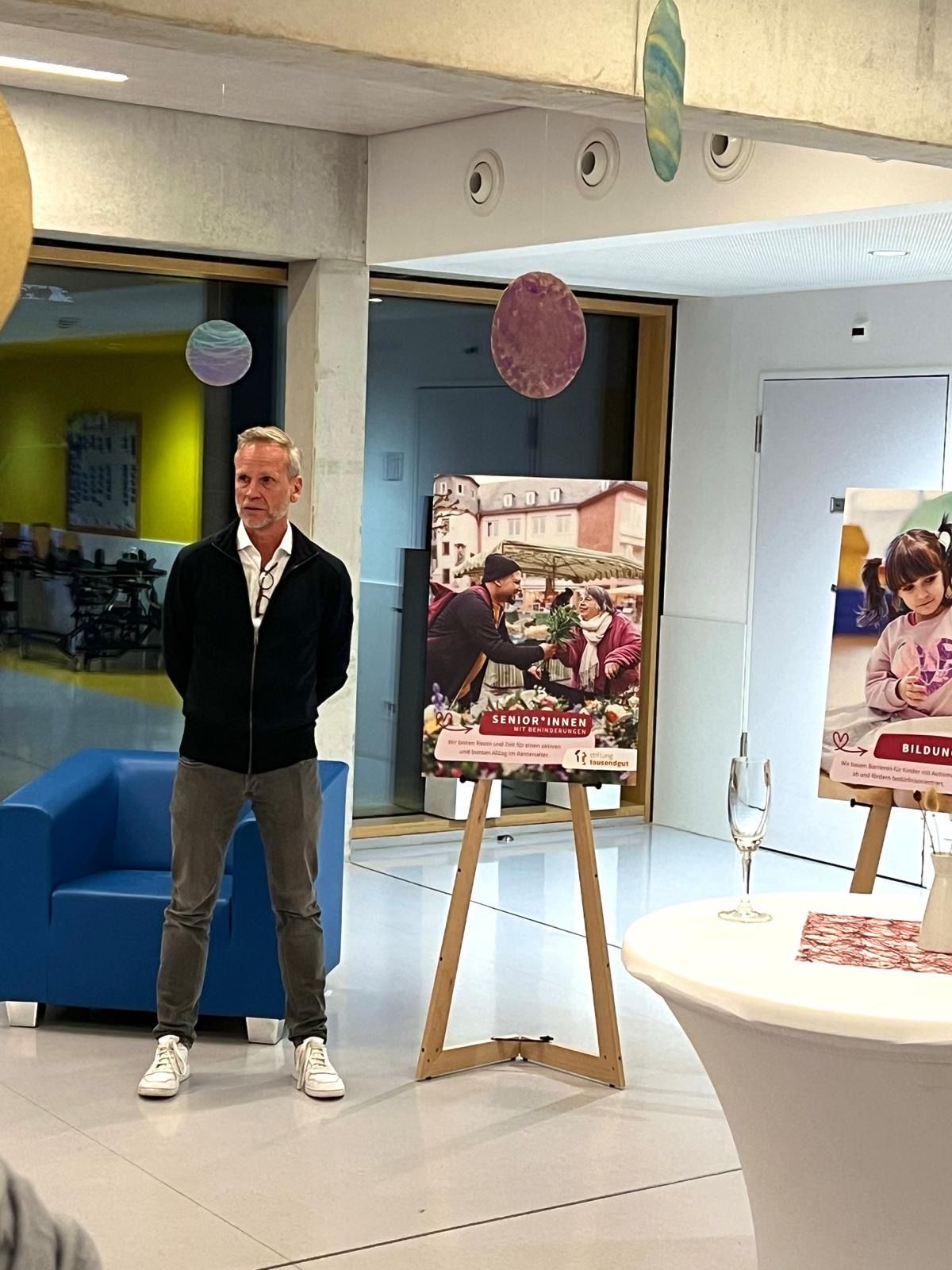 Mann steht vor einem Poster auf einer hölzernen Staffelei in einem hell erleuchteten Gebäude.