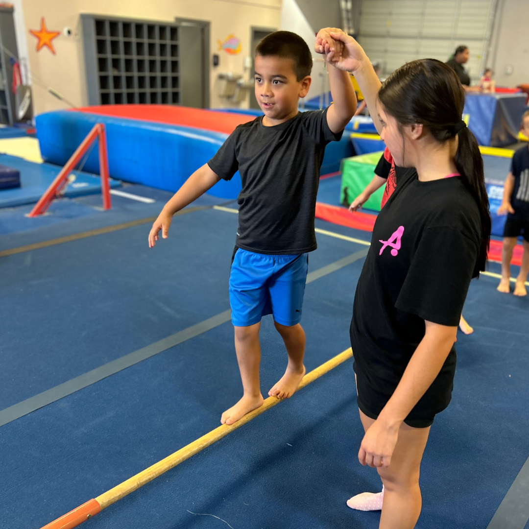 A girl in a black shirt with the letter a on it stands next to a boy on a balance beam