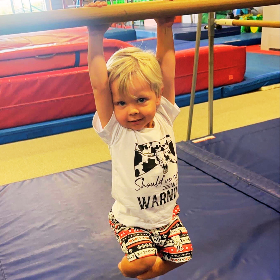 A young boy wearing a shirt that says warning is hanging from a bar