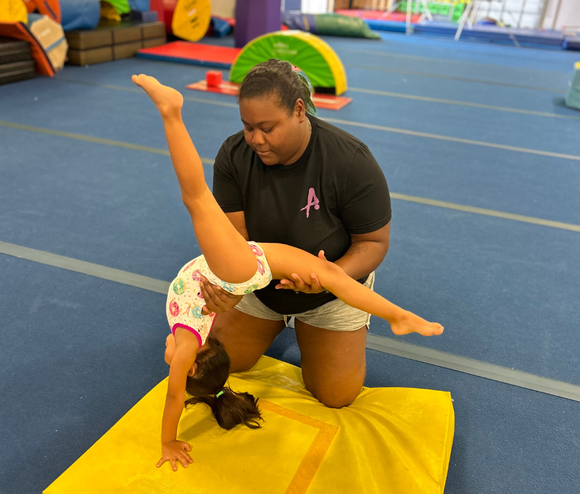 A woman is helping a young girl do a handstand on a yellow mat