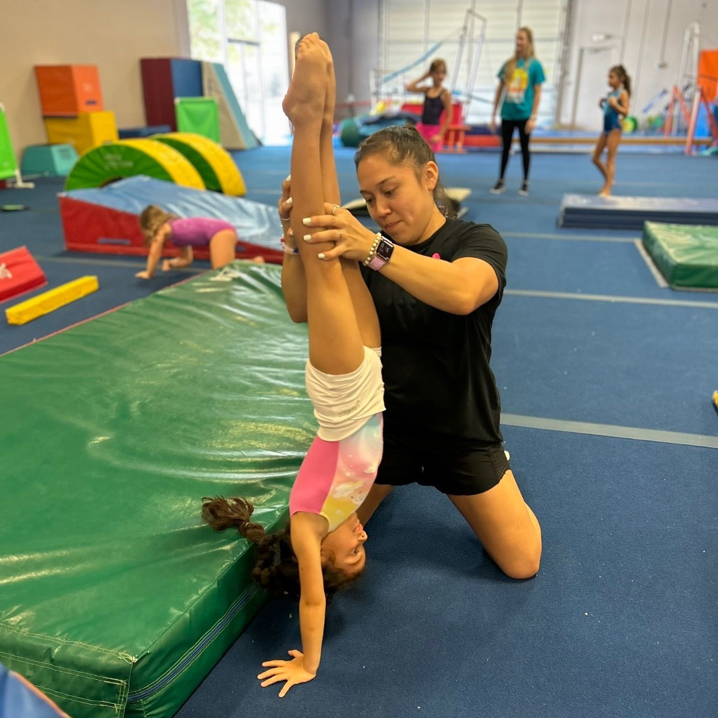 A woman is helping a young girl do a trick on an orange and blue mat that says sa sport