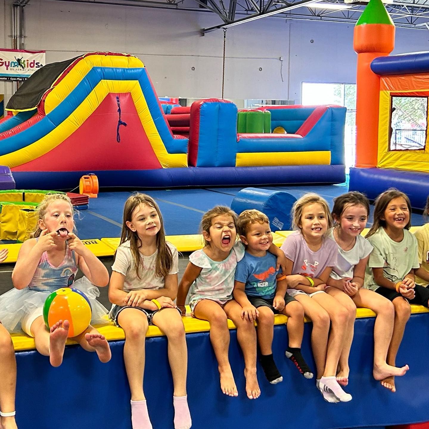 A group of children are sitting on a bench in front of a bouncy house.