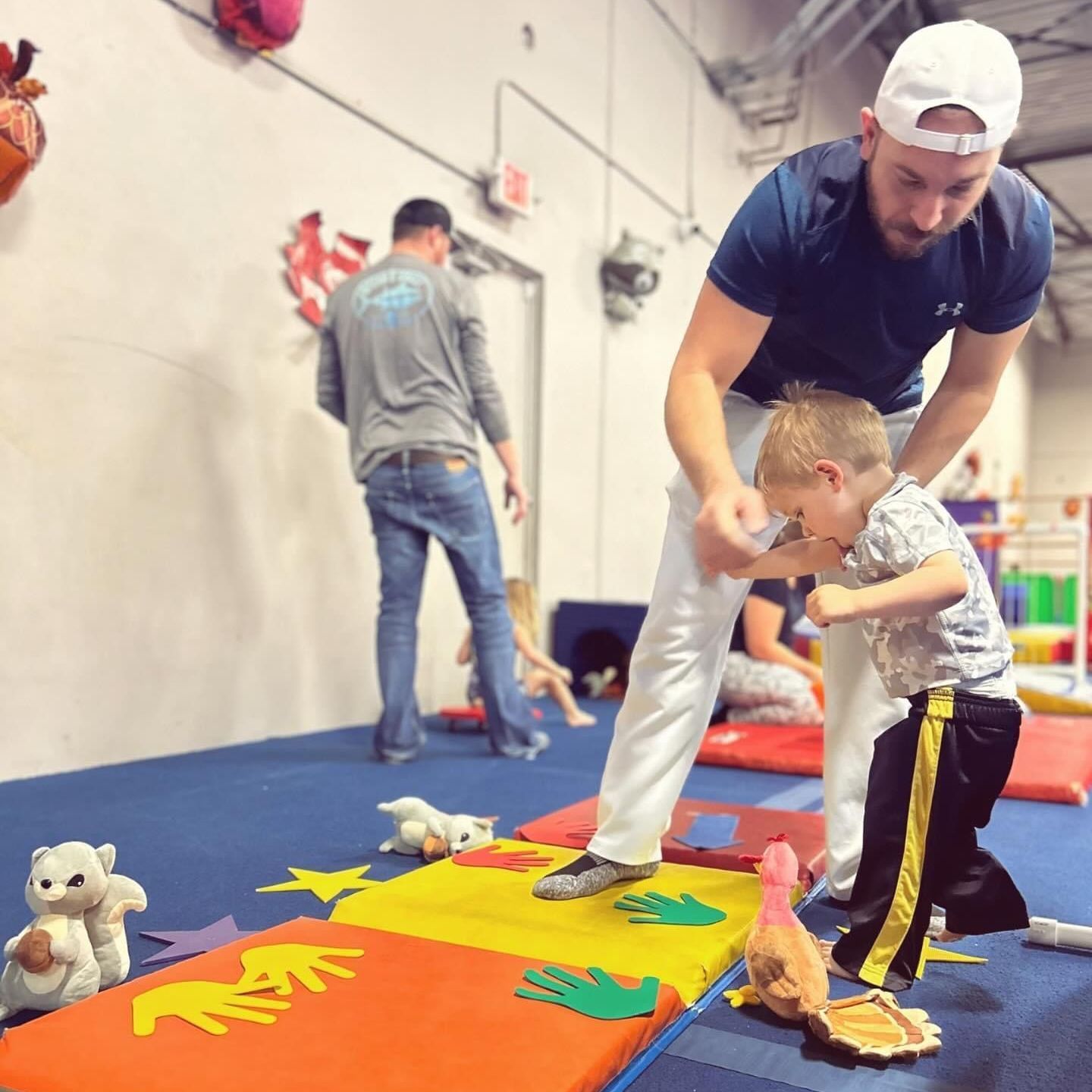 A man is helping a young boy walk on a mat in a gym.