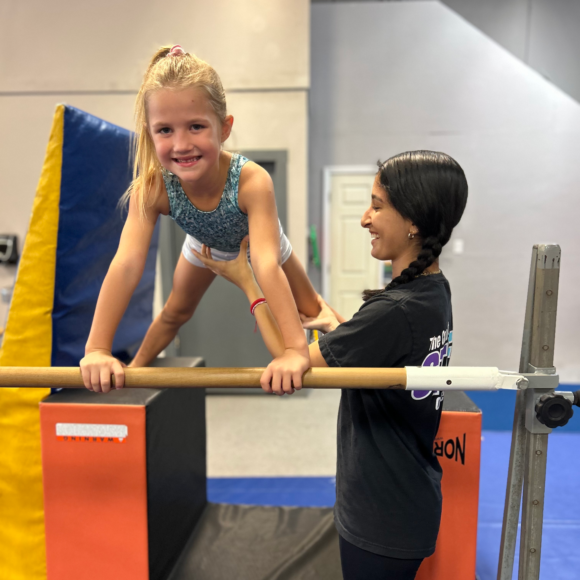 Two girls are doing a routine on a balance beam in a gym.