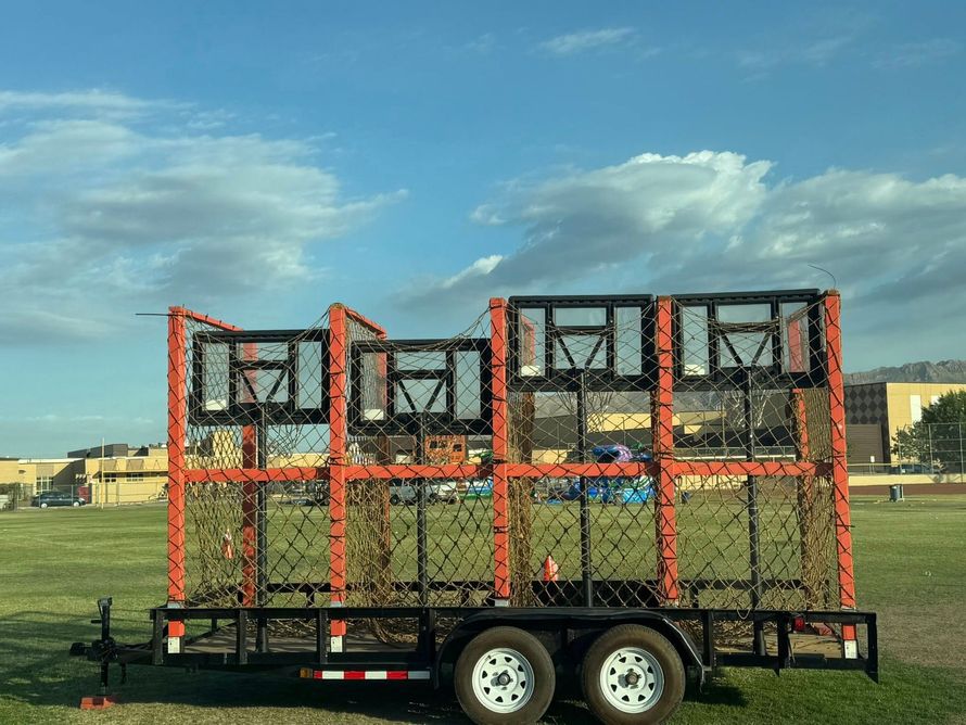 Basketball shooting game on a trailer, with orange frames and nets, set on a grassy field under a cloudy sky.