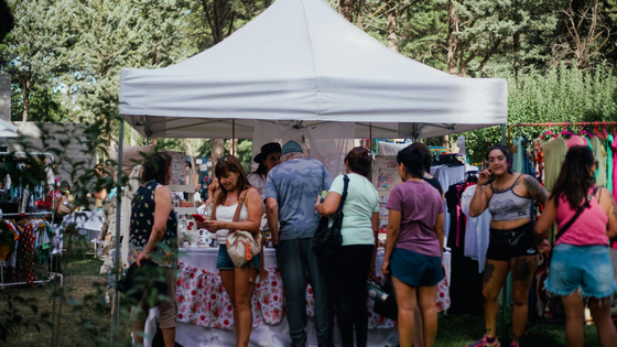People browse merchandise at an outdoor market under a white canopy tent. Some look at items, others talk.