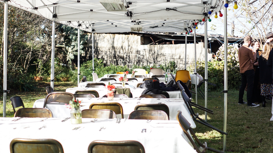 Long tables set for a meal under a white canopy in a backyard, with guests in the background.
