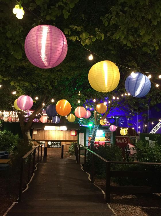 Pathway illuminated by colorful paper lanterns, leading to a venue with wooden facade, set at night.