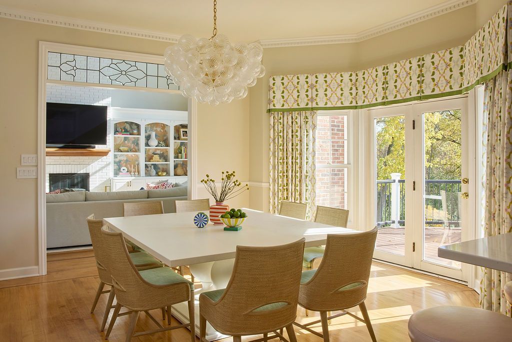 Dining room with a white square table, six wicker chairs, and a chandelier, with large arched window and outdoor view.
