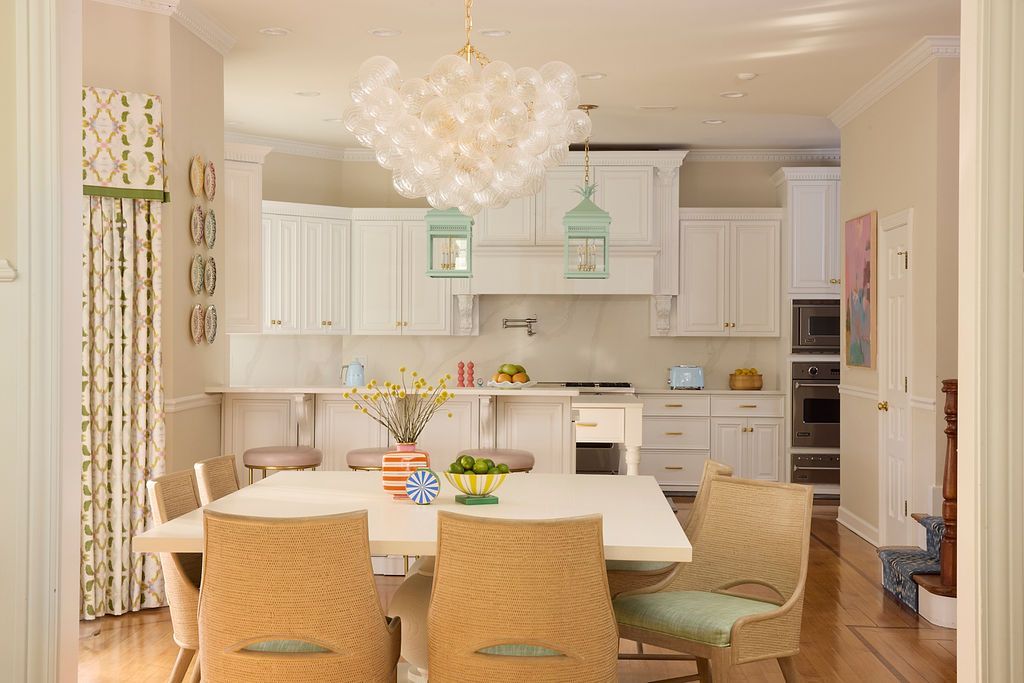 Dining room with white table, wicker chairs, and a kitchen in the background. Overhead light fixture.