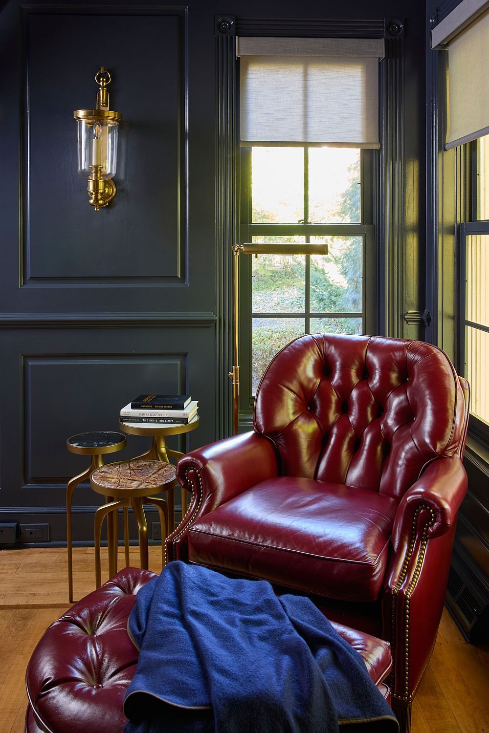Dark-paneled room with a red leather armchair and ottoman. A gold side table and lamp, with a window and a blue blanket.