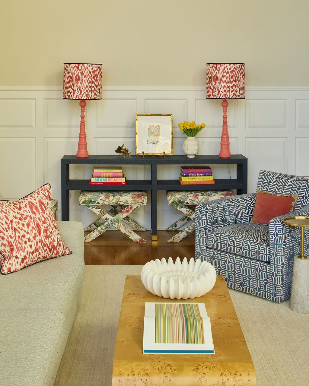 Living room with patterned armchairs, colorful lamps, and console table against a white-paneled wall.