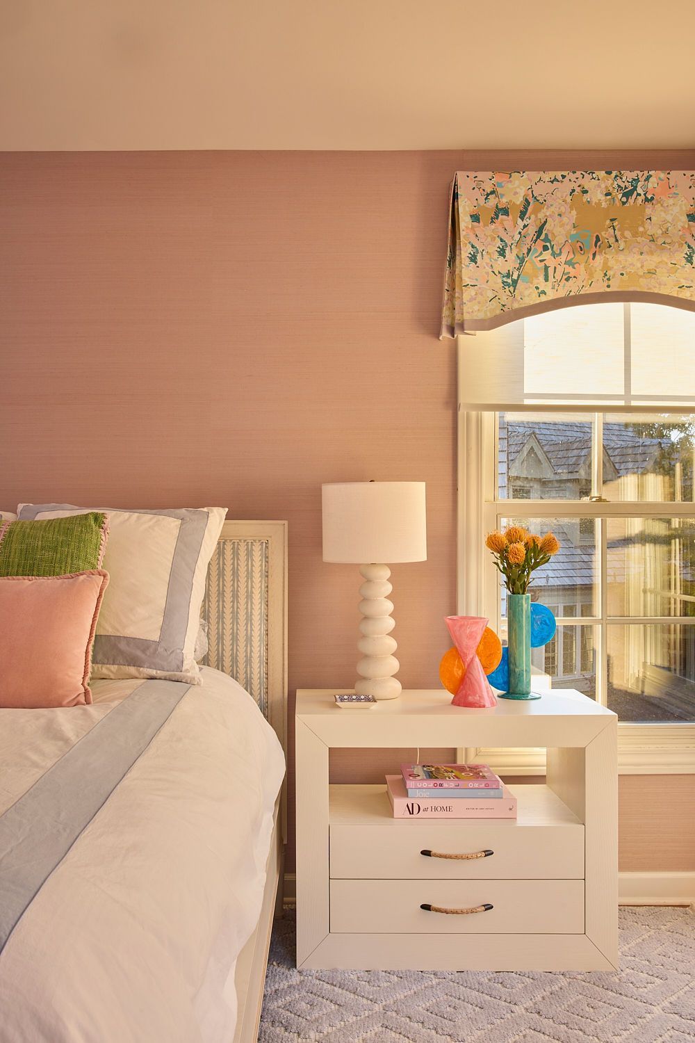 Bedroom with bed, nightstand, pink walls, window with floral valance, and colorful decor.