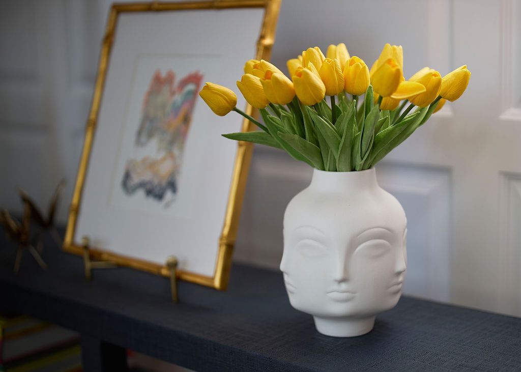 Yellow tulips in a white face-shaped vase on a blue shelf, with framed artwork in the background.