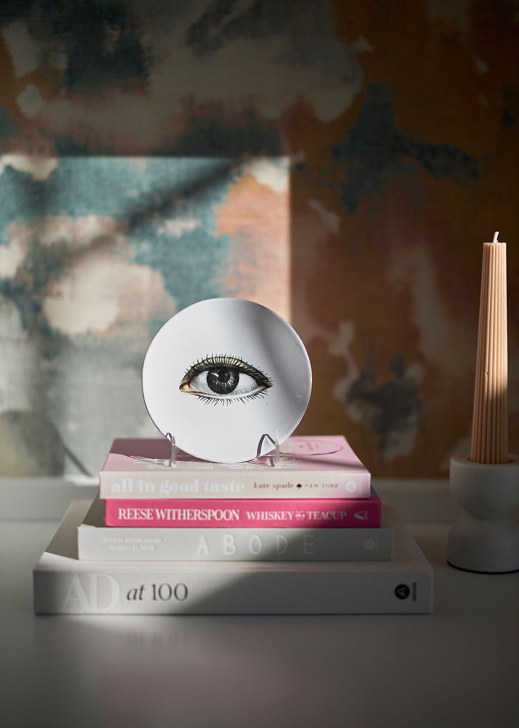 Eye illustration on white disc atop stacked books; lit candle in soft light.