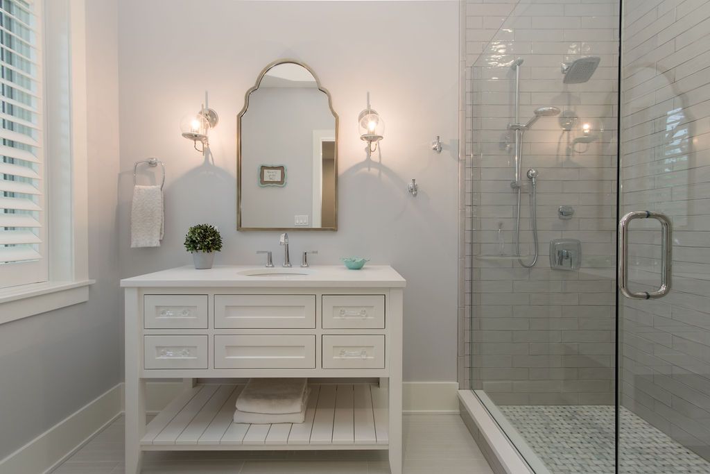 White bathroom with vanity, arched mirror, glass shower, and window with blinds.