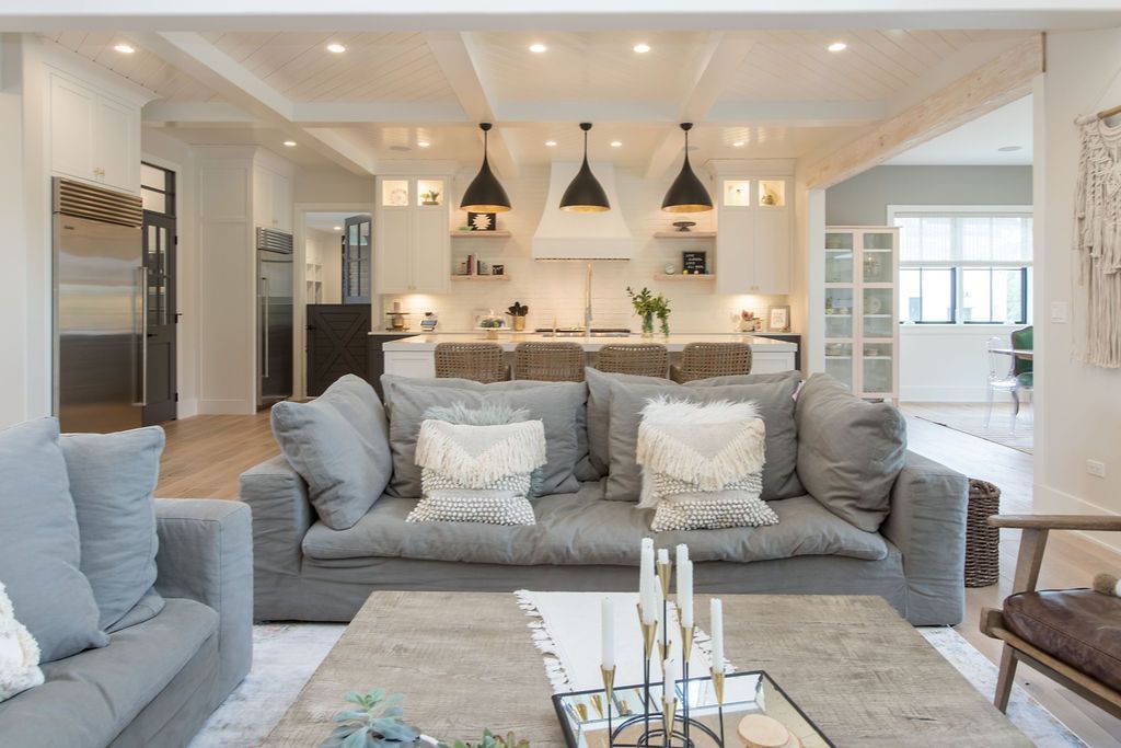 Living room with gray sofas, wooden coffee table, and kitchen in background. White walls, pendant lights.