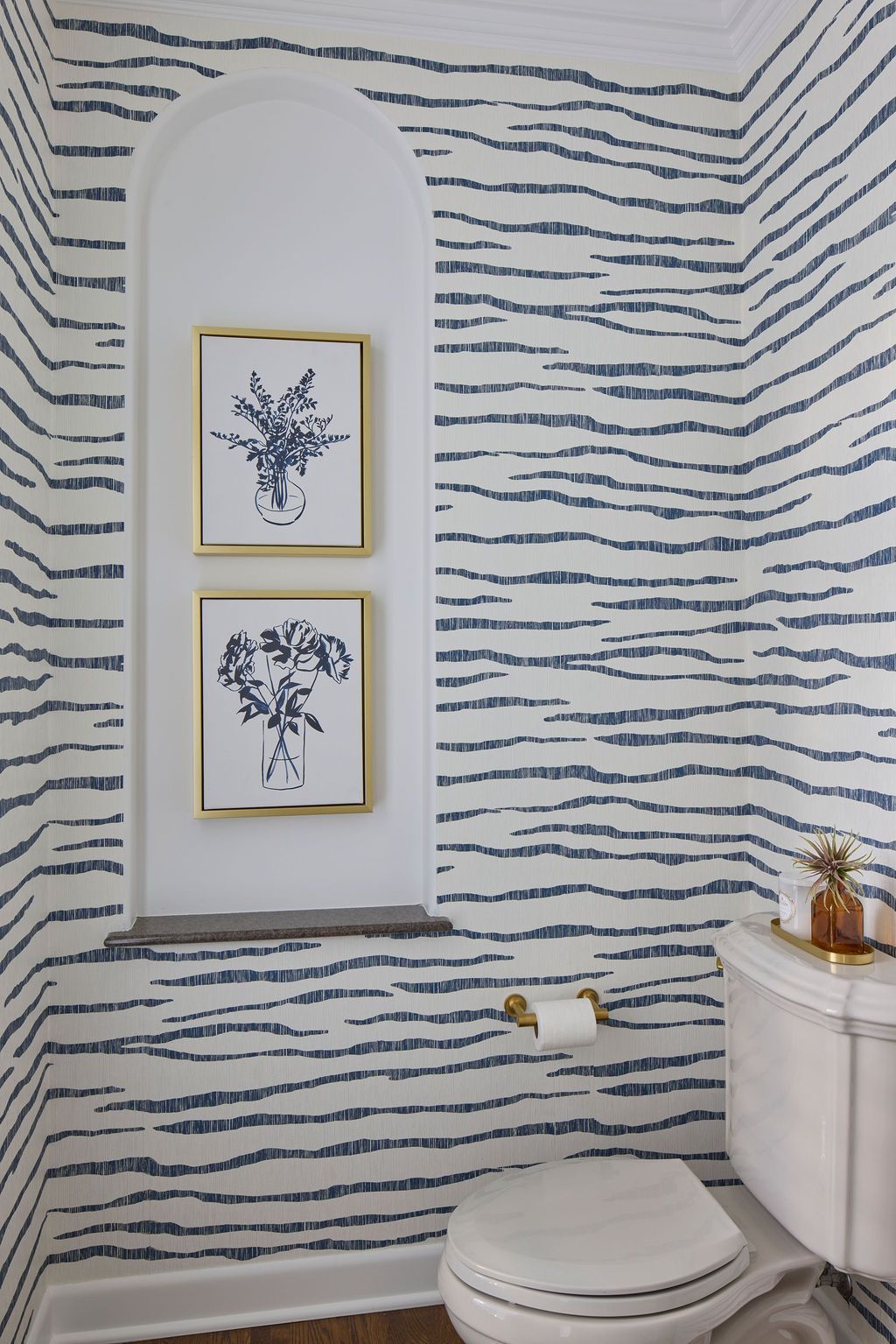 Powder room with blue and white striped wallpaper, arched alcove holding two framed botanical prints, and a white toilet.