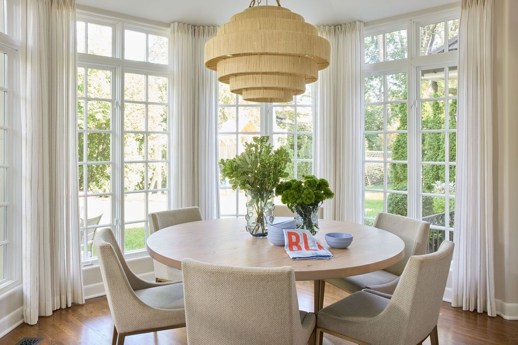 Dining room with round table, six upholstered chairs, chandelier, and large windows with white curtains.