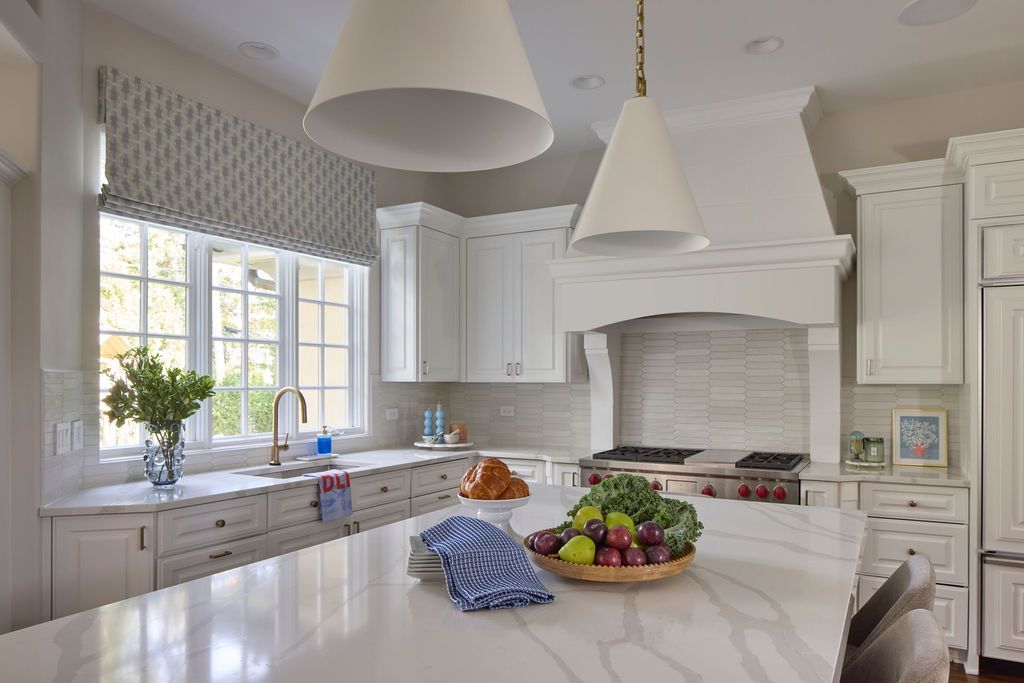 Bright, white kitchen with island, range, and cabinets. Produce in a bowl on island. Windows with patterned valance.